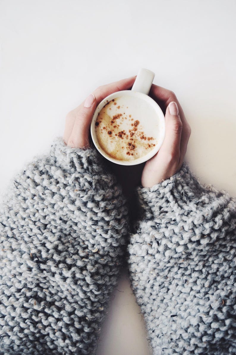 May include: A white ceramic mug filled with a creamy coffee beverage topped with a light dusting of brown cinnamon powder. The mug is held by two arms wearing a thick, chunky-knit sweater in a heathered light gray wool yarn with visible texture.