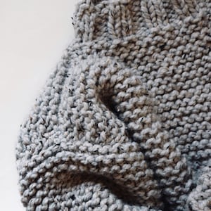 May include: A hand-knit sweater in a light gray speckled yarn featuring a ribbed neckline and a chunky garter stitch texture. The garment displays a soft, thick construction with visible flecks of darker gray and black throughout the knit.