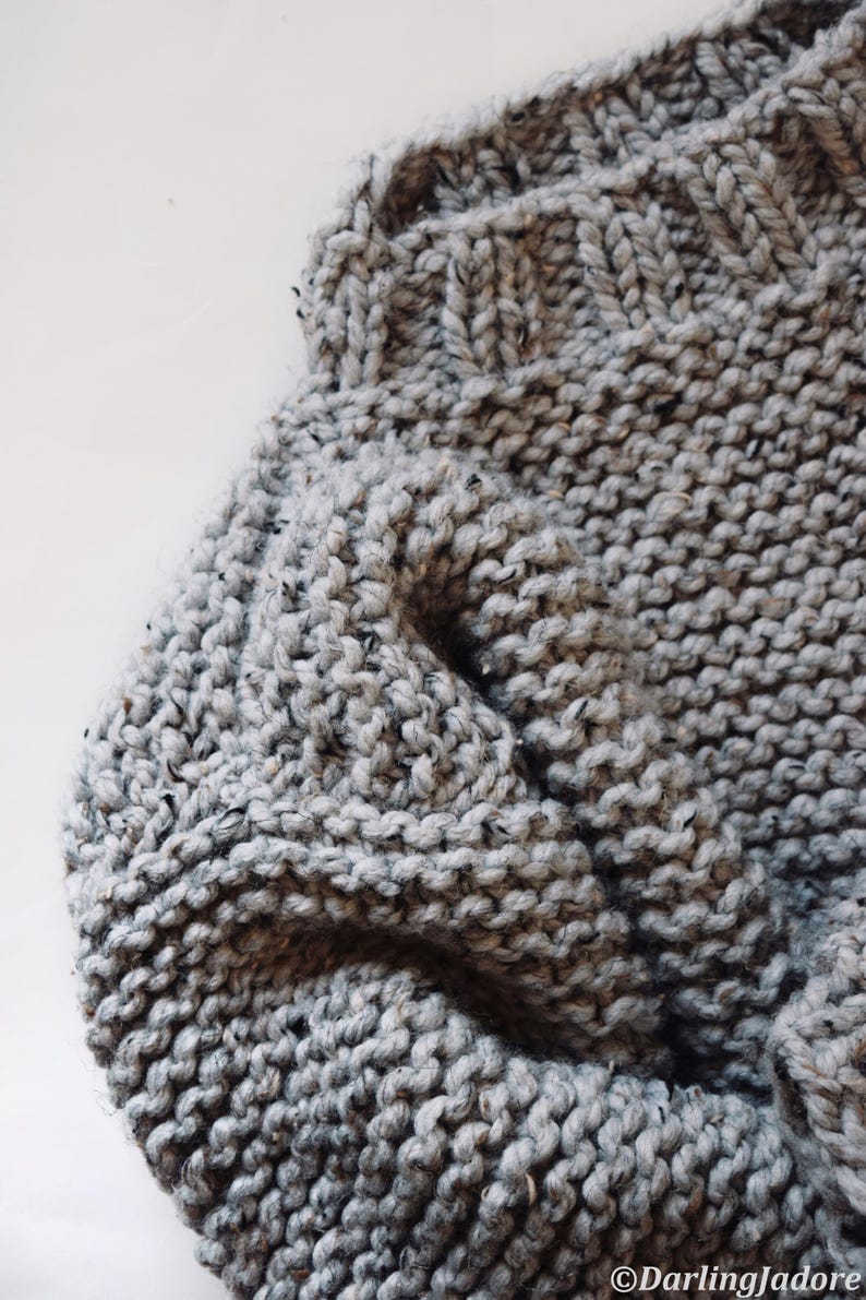 May include: A hand-knit sweater in a light gray speckled yarn featuring a ribbed neckline and a chunky garter stitch texture. The garment displays a soft, thick construction with visible flecks of darker gray and black throughout the knit.