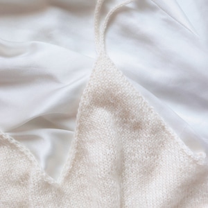 The Classic Camisole is knitted in cream yarn using this knitting pattern and and is laid flat. The spaghetti straps of this cami tank top are shown, along with a v-neck design and simple knit stitches, making it a lightweight easy camisole to knit.