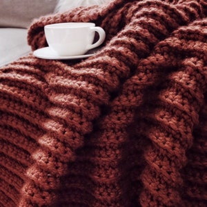 A chunky crochet blanket, The Fireside Throw, is shown crocheted in a red yarn and featuring ribbing crochet texture throughout. This chunky afghan looks warm and cozy and like it's a great blanket to crochet and use in the colder months.
