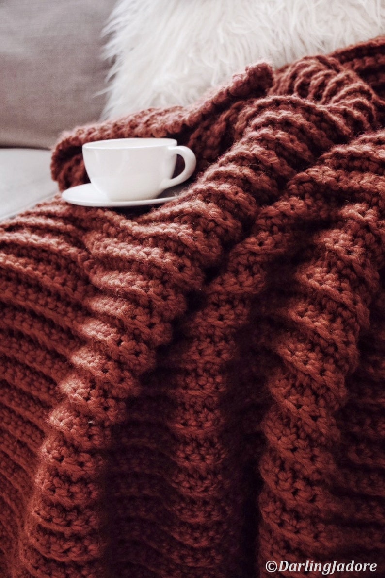 A chunky crochet blanket, The Fireside Throw, is shown crocheted in a red yarn and featuring ribbing crochet texture throughout. This chunky afghan looks warm and cozy and like it's a great blanket to crochet and use in the colder months.