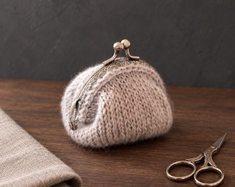 May include: A small, beige, knitted coin purse with a fuzzy texture and a vintage-style, ornate, antique brass metal kiss-lock clasp. The purse is rounded in shape and features a simple stockinette stitch pattern throughout its exterior surface.