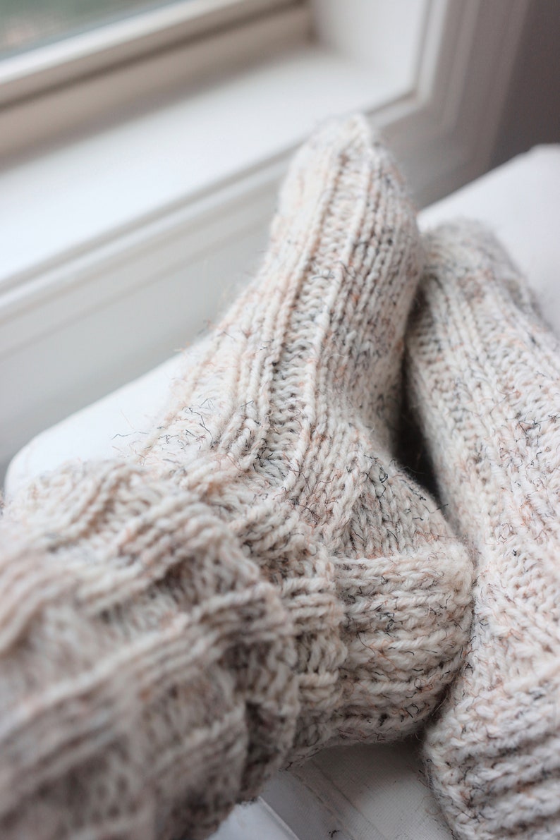 May include: A pair of thick, hand-knitted socks in a light cream color with subtle flecks of brown and grey throughout. The socks feature a prominent ribbed texture running vertically along the leg and foot, with a reinforced heel construction.