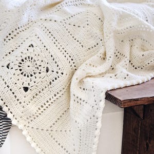 May include: An off-white crochet blanket featuring a repeating square motif pattern with intricate circular centers and a delicate bobble stitch border. The textile displays a textured, openwork design crafted with yarn, showcasing a geometric layout.
