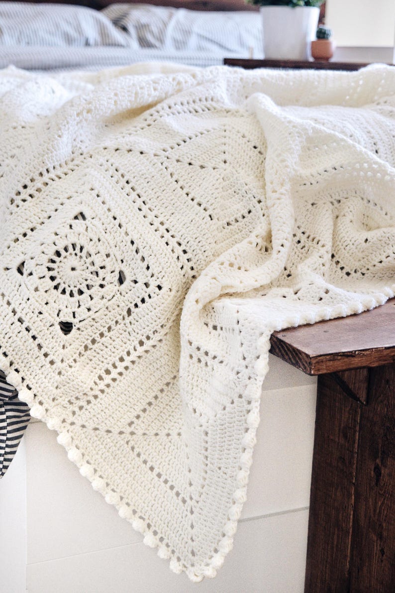 May include: An off-white crochet blanket featuring a repeating square motif pattern with intricate circular centers and a delicate bobble stitch border. The textile displays a textured, openwork design crafted with yarn, showcasing a geometric layout.