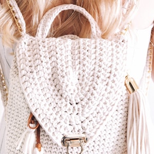 Woman wears The Lola Bag, a crochet backpack purse featuring tassels and a clasp. The bag was crocheted using a cream yarn, and there are gold metal accents for the purse hardware.  
[crochet pattern, easy crochet purse pattern, backpack purse]