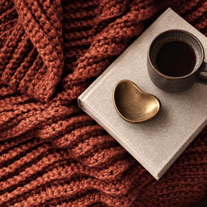 Könnte beinhalten: Eine dicke, rostfarbene Strickdecke mit strukturiertem Zopfmuster, ein hellgraues gebundenes Buch, eine kleine, glatte, herzförmige Messingschale und eine dunkle Keramiktasse mit schwarzem Kaffee auf einer dunklen Holzoberfläche.