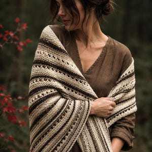 May include: A knitted shawl featuring alternating horizontal stripes of cream and dark brown yarn. The textile displays a repeating pattern of garter stitch sections interspersed with rows of eyelet lace detailing throughout the fabric.