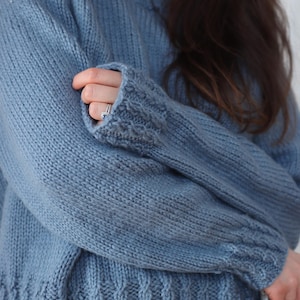 Cable knit sweater pattern, easy raglan sweater knitting pattern. women top down seamless raglan sweater with cables. oversize knit sweater pattern for women. beginner turtleneck pullover sweater knit pattern. high neck jumper knit pattern mock neck.  A light blue knitted sweater with a high neckline and textured details. The sleeves have a thumbhole design and the cuffs and hem feature a cable knit pattern. The sweater is made of a soft, possibly wool, material.