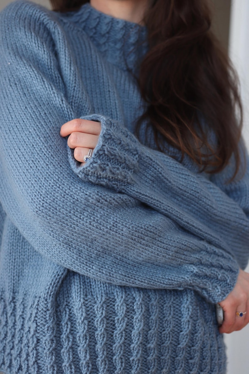 Cable knit sweater pattern, easy raglan sweater knitting pattern. women top down seamless raglan sweater with cables. oversize knit sweater pattern for women. beginner turtleneck pullover sweater knit pattern. high neck jumper knit pattern mock neck.  A light blue knitted sweater with a high neckline and textured details. The sleeves have a thumbhole design and the cuffs and hem feature a cable knit pattern. The sweater is made of a soft, possibly wool, material.