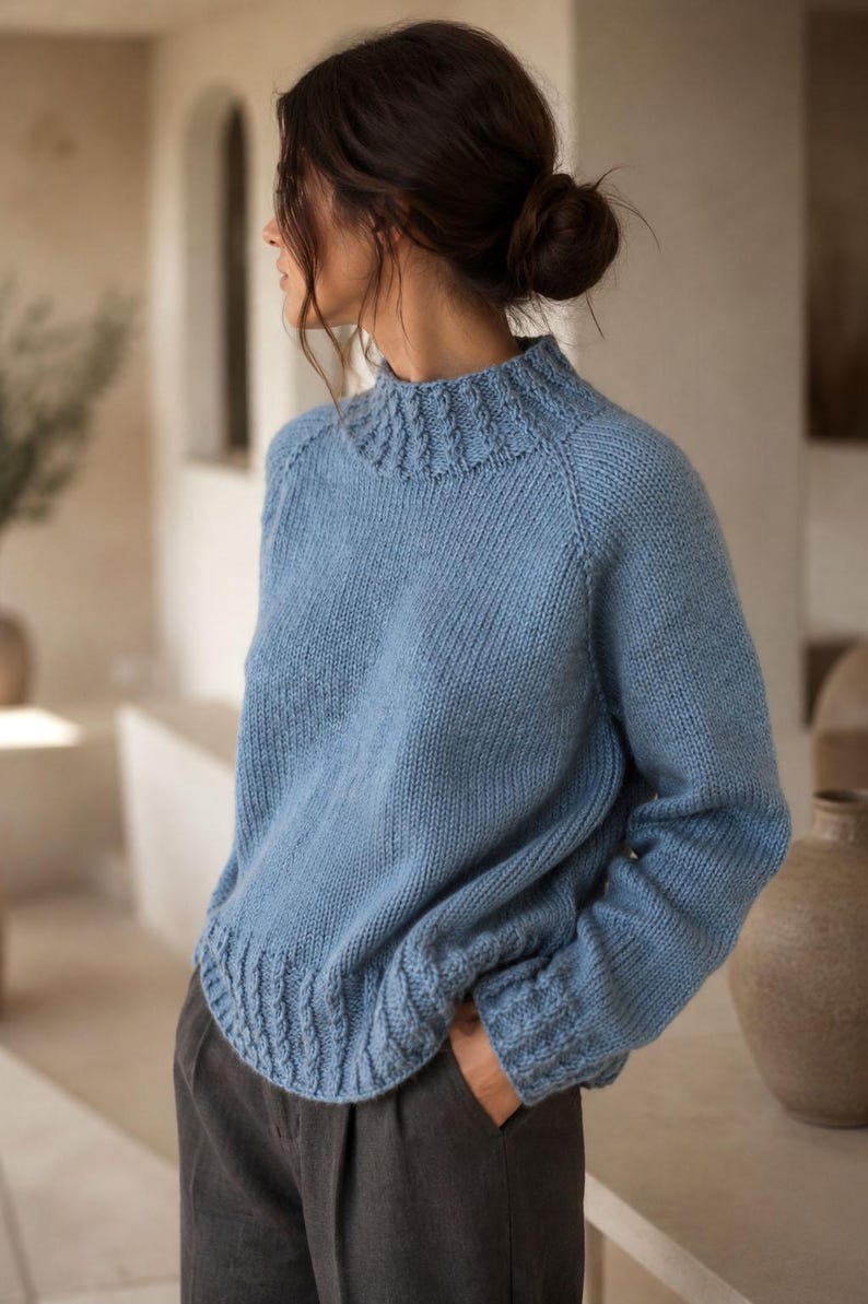Cable knit sweater pattern, easy raglan sweater knitting pattern. women top down seamless raglan sweater with cables. oversize knit sweater pattern for women. beginner turtleneck pullover sweater knit pattern. high neck jumper knit pattern mock neck.  A light blue knitted sweater with a high neckline and textured details. The sleeves have a thumbhole design and the cuffs and hem feature a cable knit pattern. The sweater is made of a soft, possibly wool, material.