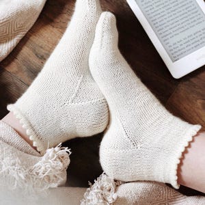 May include: Cream-coloured knitted socks with a scalloped trim, on a patterned blanket. A matching hat with a tassel and an e-reader are also visible, suggesting a cosy, relaxing setting.
