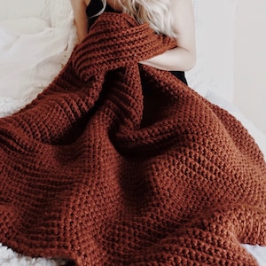 Woman holds a chunky crochet blanket, The Fireside Throw, and is using the afghan to stay warm. The blanket is crocheted in a thick red yarn and looks very warm and cozy. This easy crochet pattern is a great home decor item.
