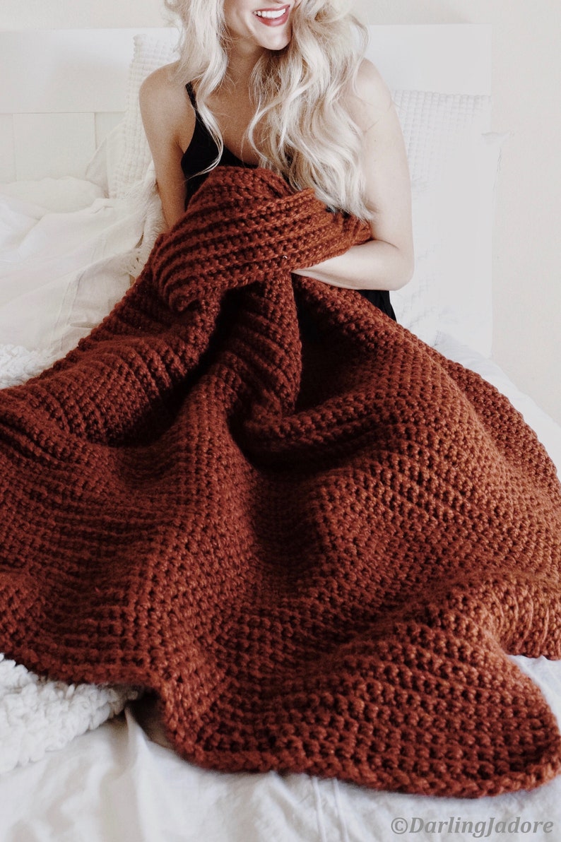 Woman holds a chunky crochet blanket, The Fireside Throw, and is using the afghan to stay warm. The blanket is crocheted in a thick red yarn and looks very warm and cozy. This easy crochet pattern is a great home decor item.