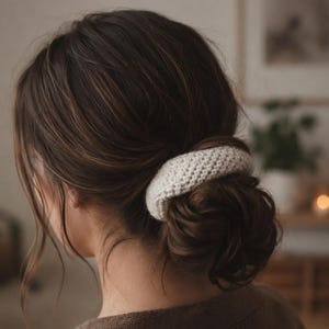 May include: A cream-colored, textured crochet hair scrunchie worn around a low bun. The accessory features a thick, woven yarn construction with a visible stitch pattern, providing a soft and rounded appearance against the dark brown hair.