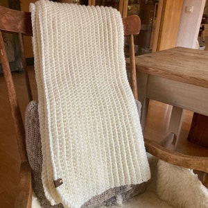 The Fireside Throw is shown crocheted in cream yarn, and it displays the ribbing texture throughout. The thick and chunky blanket looks warm and cozy and is being used as a decor item.