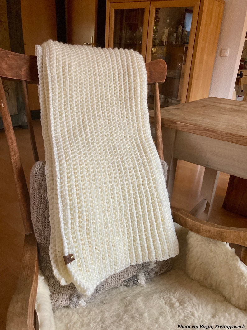 The Fireside Throw is shown crocheted in cream yarn, and it displays the ribbing texture throughout. The thick and chunky blanket looks warm and cozy and is being used as a decor item.