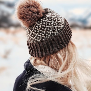May include: A dark brown and cream knit beanie featuring a geometric diamond pattern, a wide ribbed cuff, and a large, fluffy brown faux fur pom-pom on top. The hat is constructed with a thick, textured knit fabric and sits securely on the head.