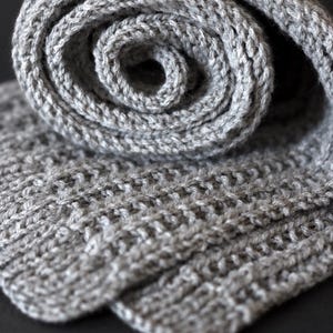 May include: A light gray knitted scarf featuring a textured ribbed pattern. The scarf is rolled into a spiral shape at the top, revealing the consistent knit structure and the soft, heathered appearance of the yarn throughout the entire piece.