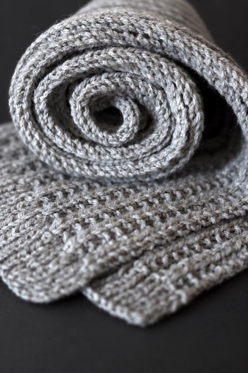 May include: A light gray knitted scarf featuring a textured ribbed pattern. The scarf is rolled into a spiral shape at the top, revealing the consistent knit structure and the soft, heathered appearance of the yarn throughout the entire piece.