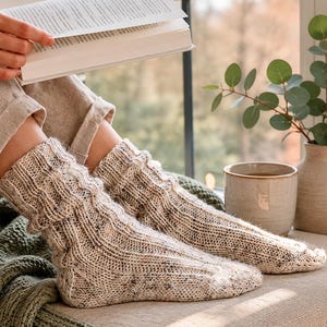 May include: A pair of cream-colored, speckled knit socks featuring a vertical ribbed texture. The socks are crafted from a textured yarn with small dark flecks throughout, extending from the ankle to the toe with a thick, cozy knit construction.