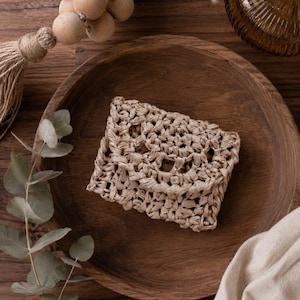 May include: A small, rectangular, beige crocheted pouch with a flap closure, featuring an open-weave texture made from natural-toned fibers. The item is centered, showcasing a consistent knotted pattern throughout its compact, envelope-like design.