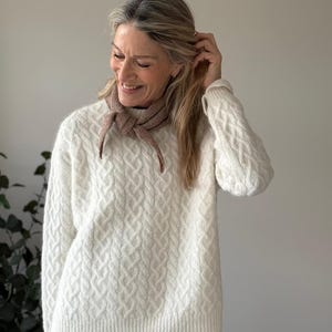 Puede incluir: Un su&eacute;ter de manga larga color crema con un patr&oacute;n de tejido de cable texturizado, un dobladillo acanalado y un cuello redondo acanalado. Una peque&ntilde;a bufanda de punto triangular de color marr&oacute;n claro est&aacute; atada al cuello. La prenda es suave.