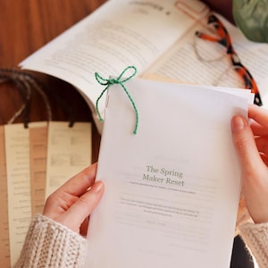 Könnte beinhalten: Eine weiße Broschüre mit dem Titel The Spring Maker Reset und einer kleinen grünen Schnur, die oben links zu einer Schleife gebunden ist. Die Broschüre enthält gedruckten Text und wird von Händen in einem beigen Strickpullover gehalten.