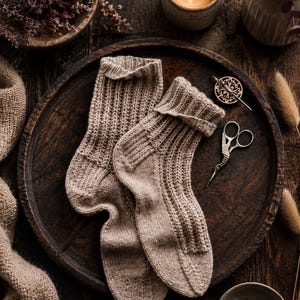 May include: A pair of light beige knit socks featuring a ribbed texture on the cuffs and legs, with a smooth knit foot section. The socks are displayed on a dark wooden tray alongside a pair of small metal scissors and a circular wooden button.
