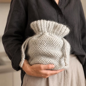 May include: A light gray, fuzzy, knitted drawstring pouch with a textured, honeycomb-like stitch pattern on the main body and a ribbed, elasticized opening at the top. The bag features two matching knitted cords for closure and carrying.