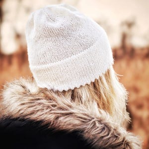 Könnte beinhalten: Eine weiße Strickmütze mit umgeschlagenem Bündchen und einfachem glatt rechts gestricktem Muster. Die Mütze wird über blondem Haar getragen und mit einer schwarzen Jacke kombiniert, die einen dicken, braunen Kunstfellbesatz am Kragen hat.
