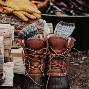 Knitted socks are inside winter hiking boots next to a fire. These knit socks, The Camp Socks, are warm and soft and look great to wear during fall or winter. 
[socks knitting pattern, knit sock patterns, easy knitting patterns, knit patterns]