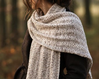 Può includere: Una lunga sciarpa rettangolare in maglia di filato screziato beige chiaro e crema. La sciarpa presenta un motivo a trama ripetitiva con chevron o zigzag verticali lungo tutta la lunghezza, drappeggiata sulle spalle sopra un cappotto scuro.