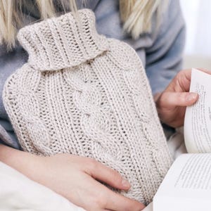 Könnte beinhalten: Eine beige Strickhülle für eine Wärmflasche mit Zopfmuster und Rollkragen. Die Hülle besteht aus einem weichen, strukturierten Garn. Die Wärmflaschenhülle soll Wärme und Komfort bieten.