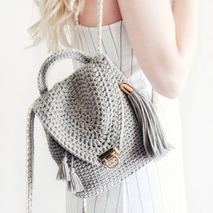 Woman holds The Lola Bag, a crocheted backpack purse over one shoulder, with one crochet strap hanging down. This crochet backpack was crocheted using gray yarn, and features tassels and hardware accents. 
[crochet pattern, crochet backpack]