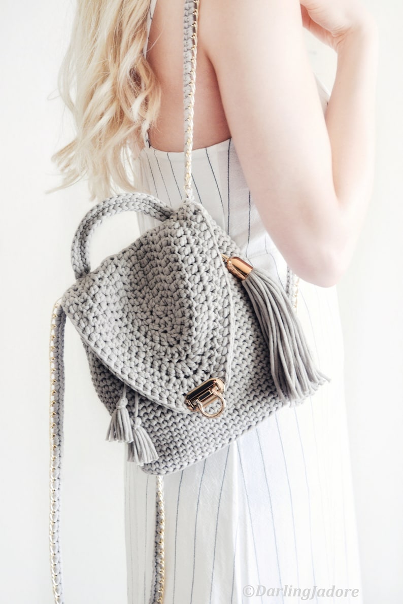 Woman holds The Lola Bag, a crocheted backpack purse over one shoulder, with one crochet strap hanging down. This crochet backpack was crocheted using gray yarn, and features tassels and hardware accents. 
[crochet pattern, crochet backpack]