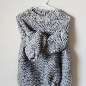 May include: A chunky knit sweater in a light gray color with a speckled texture. The sweater features long sleeves, a ribbed crew neckline, and a ribbed hem. It is displayed on a white plastic hanger, showcasing the thick yarn construction.