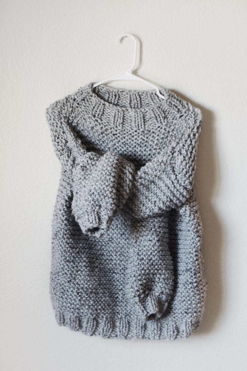 May include: A chunky knit sweater in a light gray color with a speckled texture. The sweater features long sleeves, a ribbed crew neckline, and a ribbed hem. It is displayed on a white plastic hanger, showcasing the thick yarn construction.