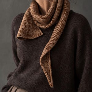 Può includere: Un maglione in maglia a maniche lunghe marrone scuro abbinato a una sciarpa in maglia a legaccio marrone chiaro. La sciarpa presenta un'estremità affusolata e appuntita ed è avvolta attorno al collo. Il maglione ha un semplice girocollo.