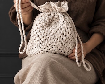 Puede incluir: Un bolso con cordón de color crema elaborado con un patrón de ganchillo de cuadrados de la abuela. El bolso presenta un cierre superior fruncido, asegurado por dos largas correas de cuerda trenzada que permiten abrir y cerrar el compartimento.