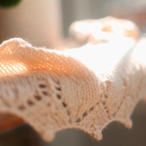 May include: Close-up of a light peach-colored knitted shawl. The shawl features a delicate openwork pattern along the edge. The soft texture and color suggest a cozy, handmade item, perfect for adding warmth and style.