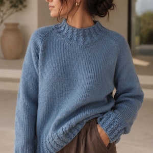 Cable knit sweater pattern, easy raglan sweater knitting pattern. women top down seamless raglan sweater with cables. oversize knit sweater pattern for women. beginner turtleneck pullover sweater knit pattern. high neck jumper knit pattern mock neck.  A light blue knitted sweater with a high neckline and textured details. The sleeves have a thumbhole design and the cuffs and hem feature a cable knit pattern. The sweater is made of a soft, possibly wool, material.