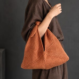 Peut inclure: Un sac en tricot de couleur rouille, de forme triangulaire, présentant un motif de mailles texturé. Le sac possède une poignée nouée sur le dessus, créant une silhouette géométrique distincte avec des coutures visibles formant des panneaux.