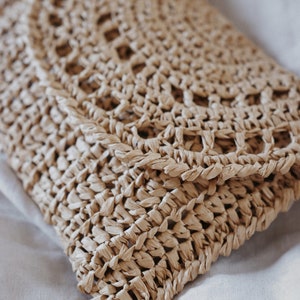 A raffia clutch purse is laid flat, knitted in raffia yarn and using the crochet pattern. This summer clutch bag is a great sized purse to hold essentials to store things. This pretty summer straw crochet clutch is easy to crochet and fashionable.