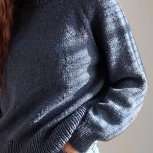 Può includere: Un maglione in maglia blu scuro a maniche lunghe con collo alto, polsini a coste e orlo a coste. Il tessuto in maglia presenta una sottile trama mélange con sfumature di blu più chiaro e grigio su tutto il capo.