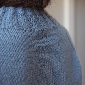 May include: A light blue knitted sweater featuring a textured cable knit pattern around the neckline and a smooth stockinette stitch throughout the body. The garment is crafted from a soft yarn with a visible wool-like texture and a consistent hue.