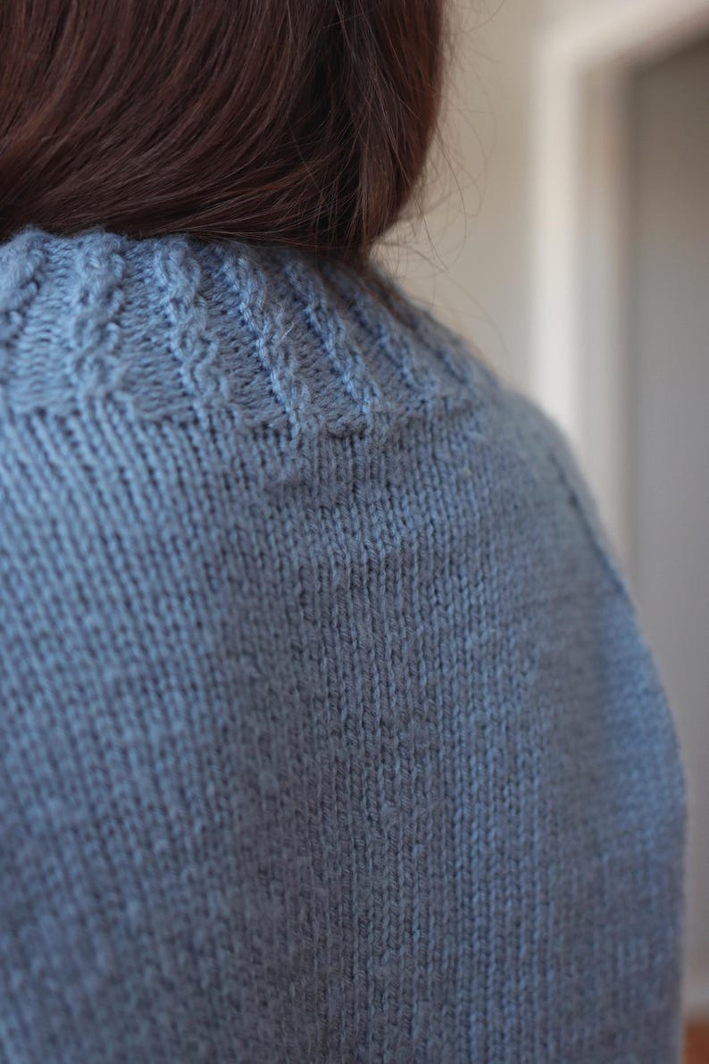 May include: A light blue knitted sweater featuring a textured cable knit pattern around the neckline and a smooth stockinette stitch throughout the body. The garment is crafted from a soft yarn with a visible wool-like texture and a consistent hue.