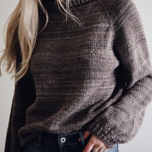 May include: A brown knit sweater with long sleeves and a crew neck. The sweater is paired with blue jeans. The image is a medium shot, focusing on the upper body and clothing. Beginner sweater knitting pattern, oversized top-down raglan pullover in worsted weight yarn, women's seamless crewneck knit jumper. ribbon tie knit sweater pattern. oversize raglan jumper pattern for women