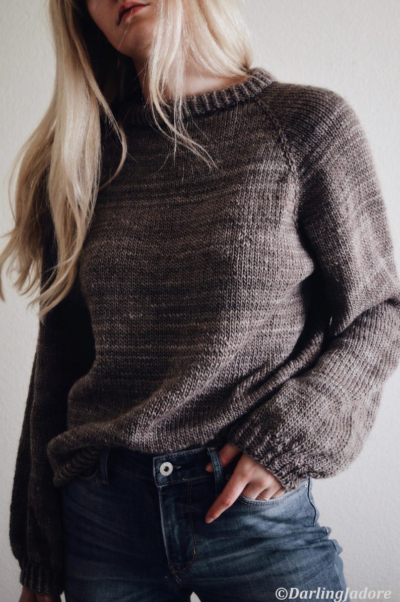 May include: A brown knit sweater with long sleeves and a crew neck. The sweater is paired with blue jeans. The image is a medium shot, focusing on the upper body and clothing. Beginner sweater knitting pattern, oversized top-down raglan pullover in worsted weight yarn, women's seamless crewneck knit jumper. ribbon tie knit sweater pattern. oversize raglan jumper pattern for women